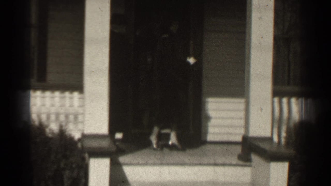 A dark-coated figure stands near a front doorway on a porch with columns and shrubs.