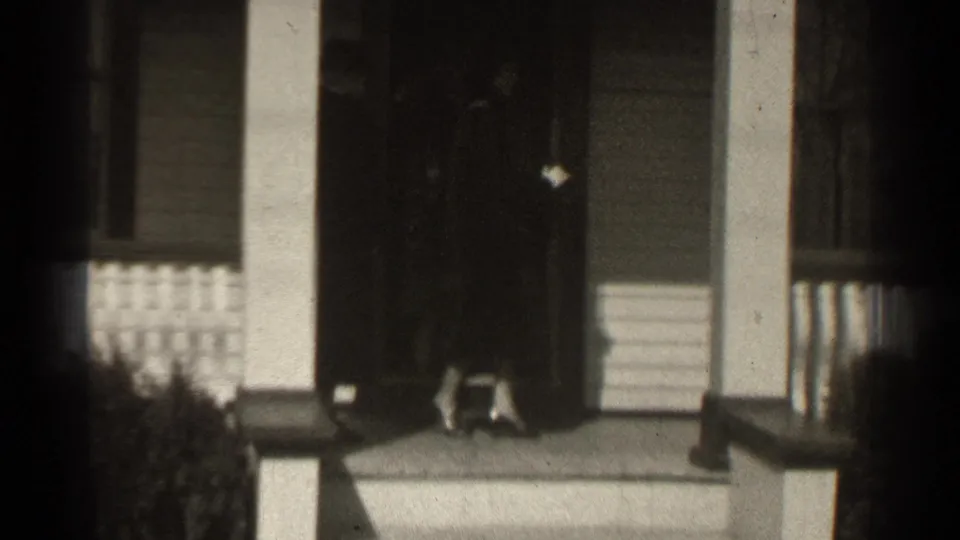 1939 New York home-movie frame showing a person in a dark coat near a residential front door and porch columns.