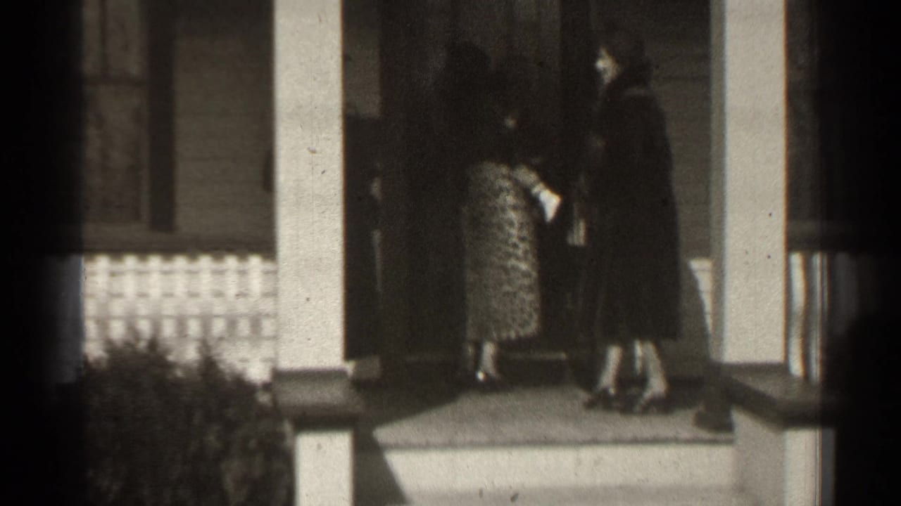 Two women and a man stand on a porch near a doorway and white columns.