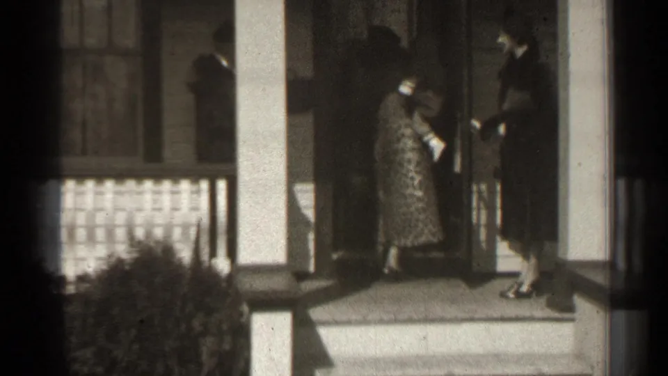 Adults are clustered at a residential doorway in 1939 New York footage, including a woman in a patterned dress holding a handbag.