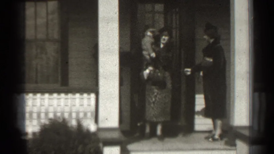 In a supplied 1939 New York home-movie frame, women gather on a columned porch as one holds a baby near the doorway.