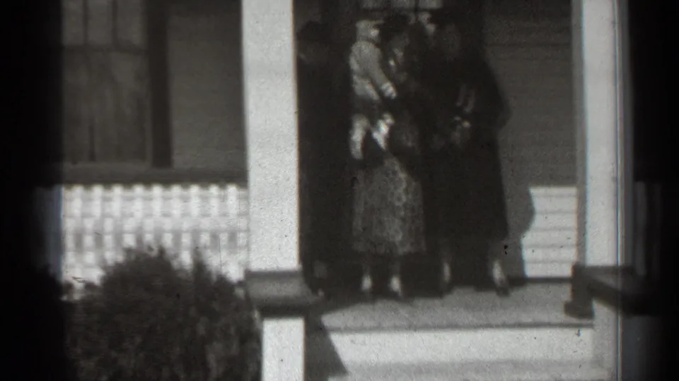 A soft 8mm frame shows women gathered tightly on a porch, with a baby held near the center of the group.