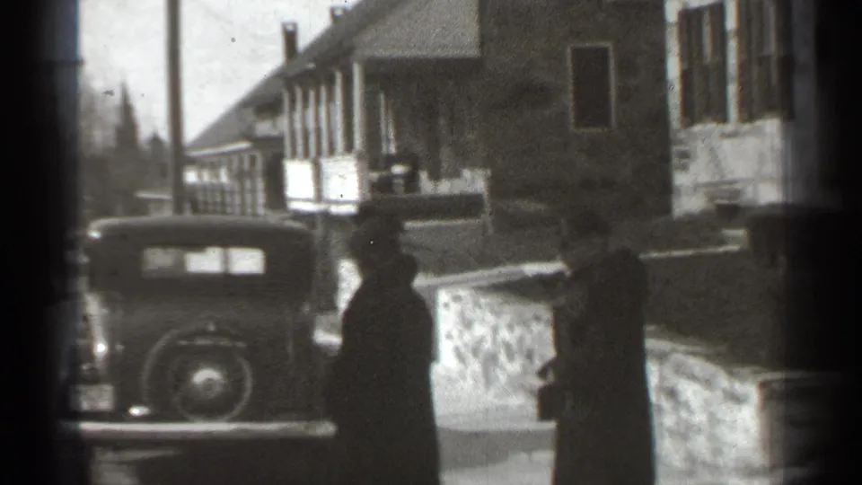 Two women in dark outerwear move near a parked prewar-style car on a residential New York street, per metadata.