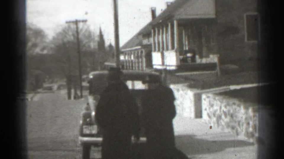 The women continue along the residential street near a parked vintage car, with bare trees and utility poles in view.