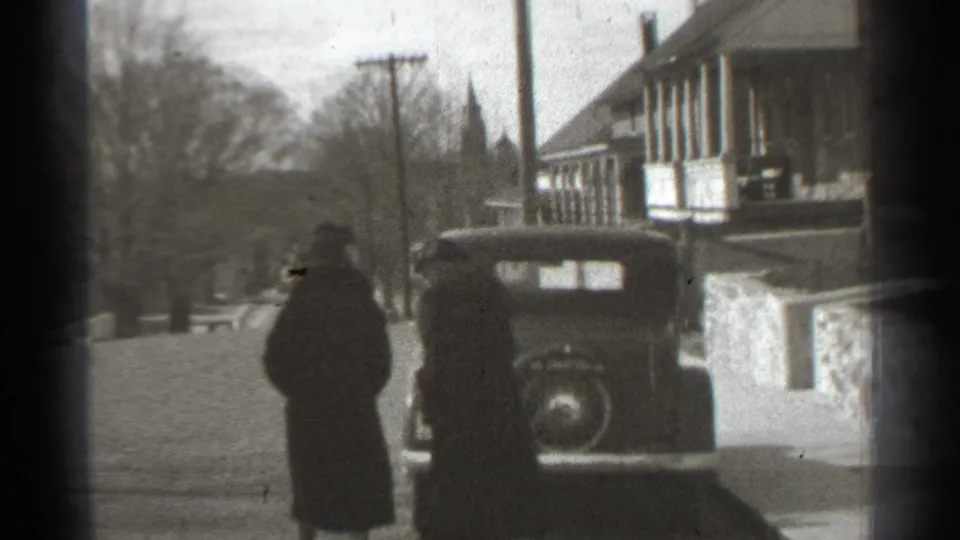 A woman in a long dark coat is seen near the parked car on the residential street, with another figure partly obscured at the edge.