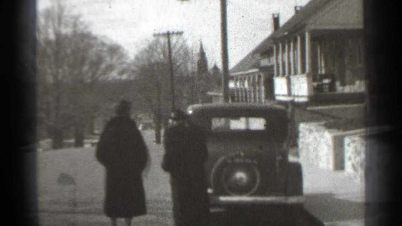 In supported 1939 New York footage, two adults in dark coats move toward a parked prewar car beside a house-lined street.