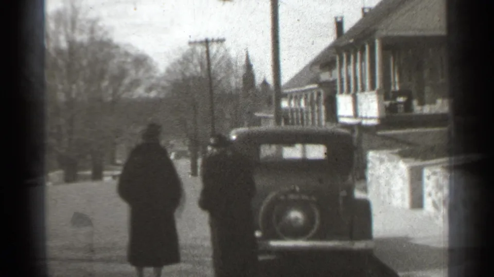 In supported 1939 New York footage, two adults in dark coats move toward a parked prewar car beside a house-lined street.