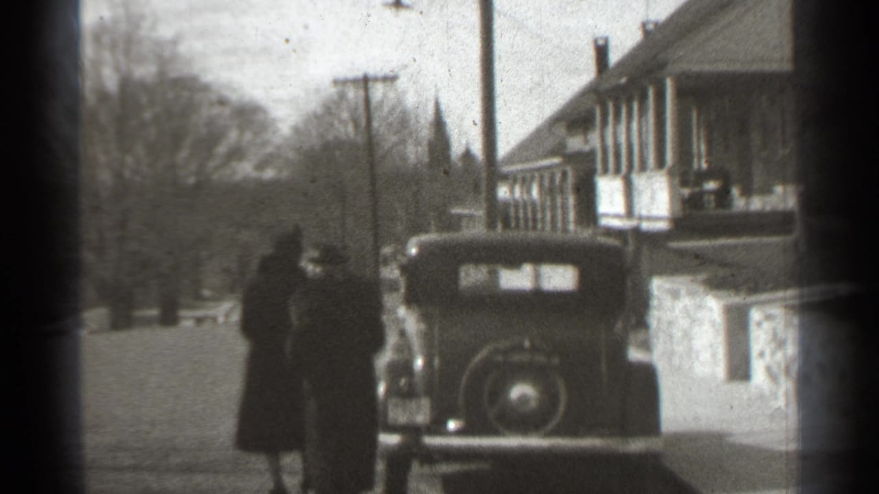 Two adults stand beside a parked vintage car near a house and utility pole.