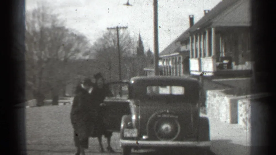 The two adults remain beside the parked prewar car, with one positioned at the door on the quiet New York street.