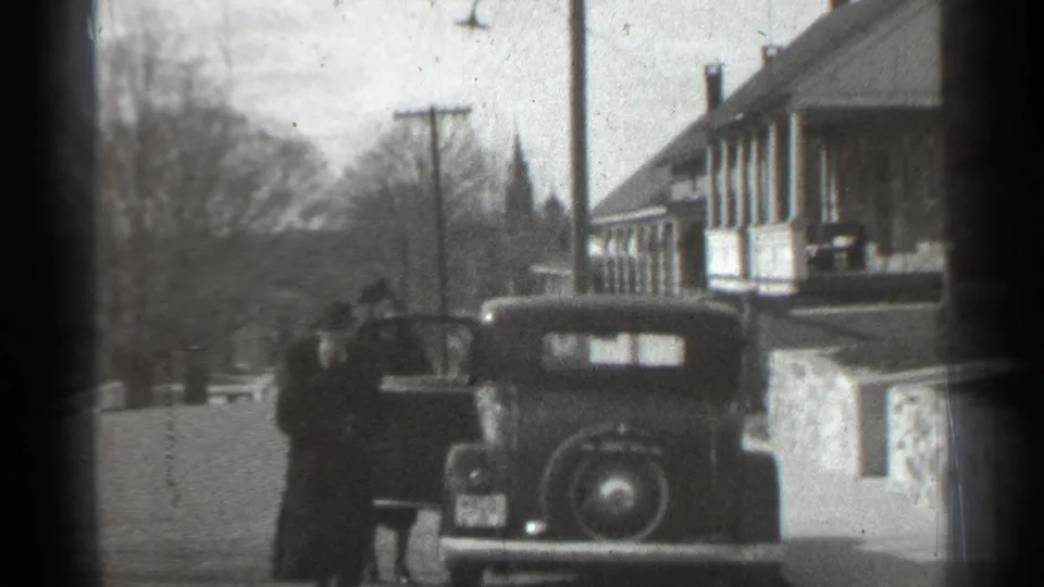 1939 New York home-movie frame showing an older car parked beside a porch-front building with one person near the open door.