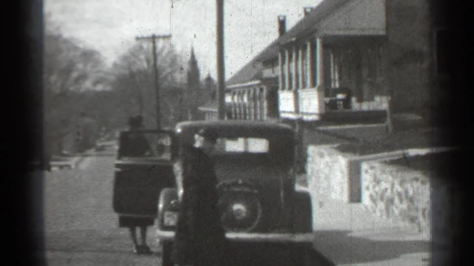 1939 New York frame with a parked older automobile, an open door, and a figure standing on the street near porch-front buildings.