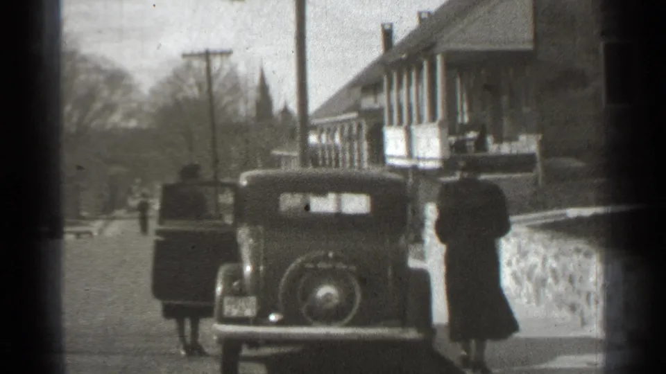 1939 New York home-movie frame showing a parked car, an open door, and two adults near a residential-looking building.