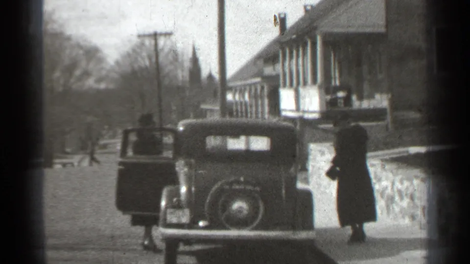 1939 New York frame with two adults in dark coats near a parked older car beside porch-front buildings and utility poles.