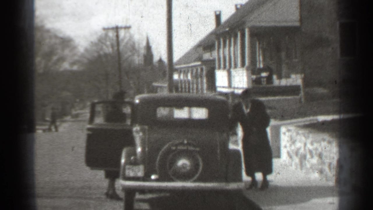 Two adults stand by a dark 1930s car with open doors on a residential street.