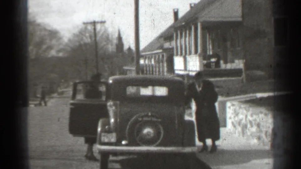 New York 1939 home-movie frame showing adults beside a dark classic car near porch-front houses and a distant steeple.