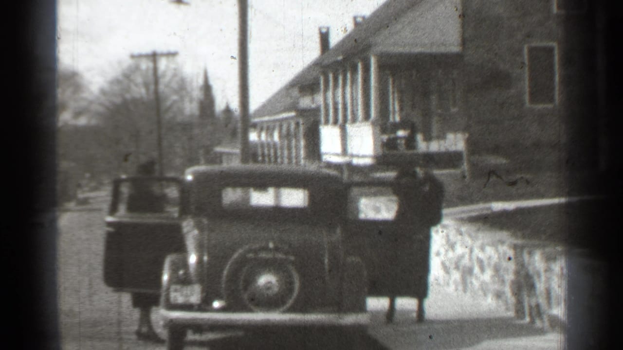 A woman stands at the rear of a dark car while another person is near the open side door.