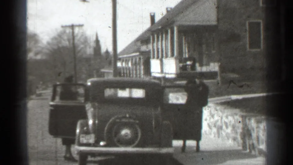New York 1939 frame with a woman in a long coat at the rear of a classic car outside residential buildings.