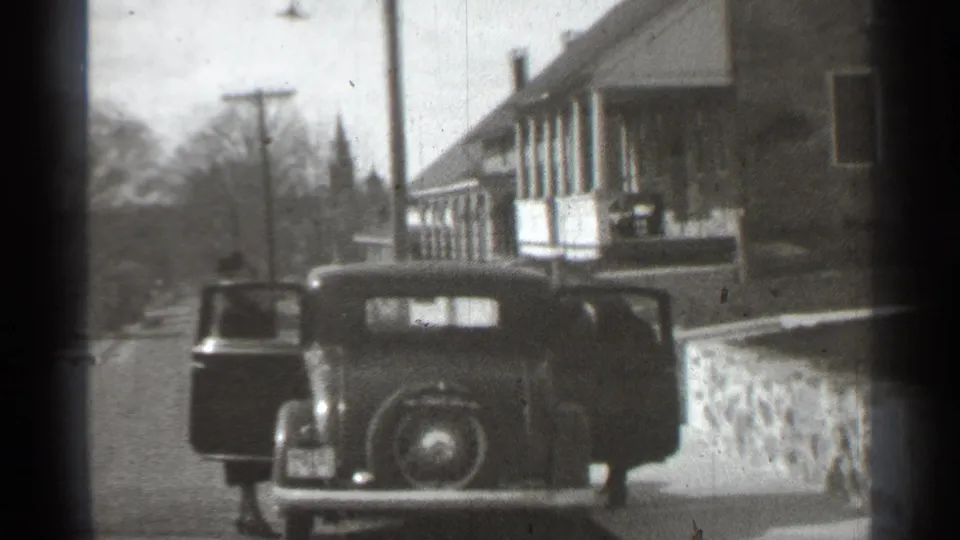 1939 New York home-movie frame showing a classic car on a suburban street with a person partly visible by the open door.