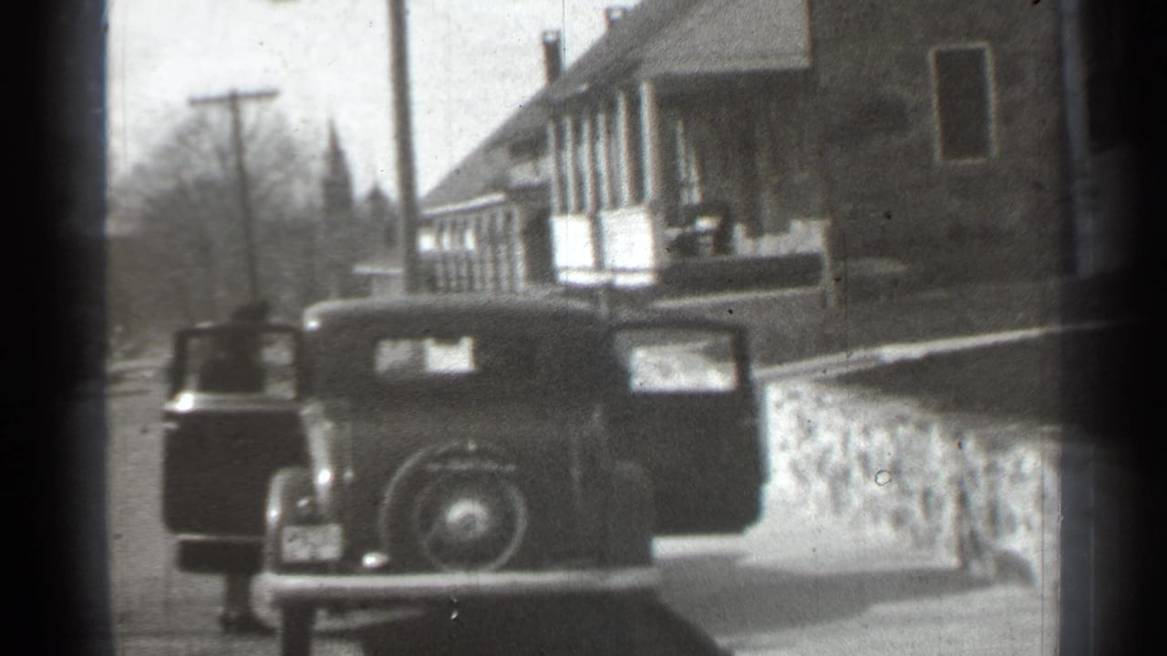 1939 New York home-movie frame showing a parked car with open doors on a residential street near a stone wall and house.