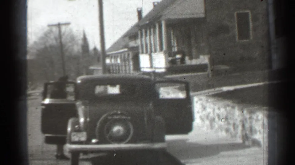 1939 New York home-movie frame showing a parked car with open doors on a residential street near a stone wall and house.
