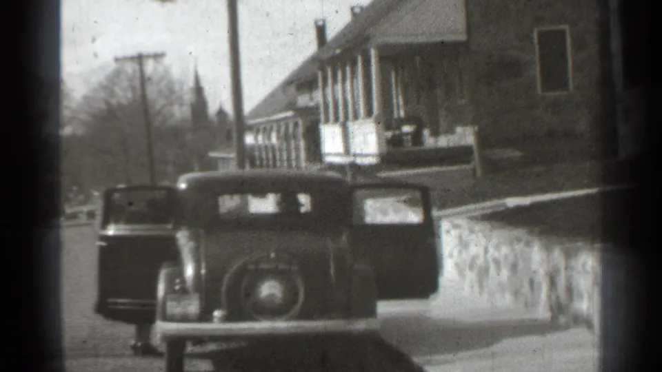 1939 New York frame of a parked automobile with open doors, a house porch, stone wall, and distant steeple in view.