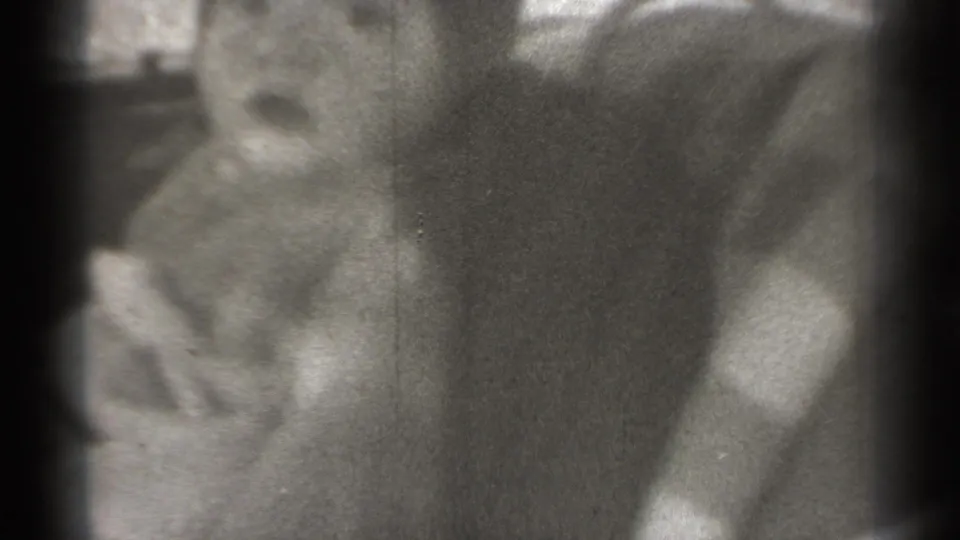 Grainy 1939 New York 8mm frame of an infant in light clothing seated in a car interior, with window light behind.