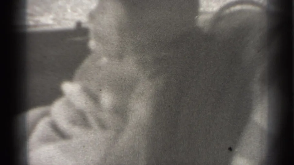 Grainy black-and-white 8mm frame of a baby seated inside a car, with soft focus and dim interior shapes around the child.
