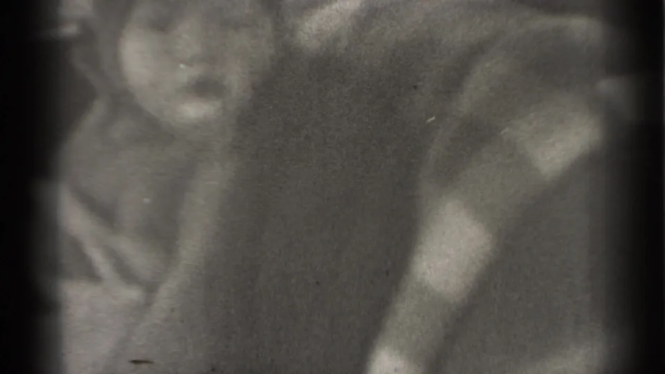 A baby turns within a vehicle-like interior while a blurred adult figure appears behind the seat area.
