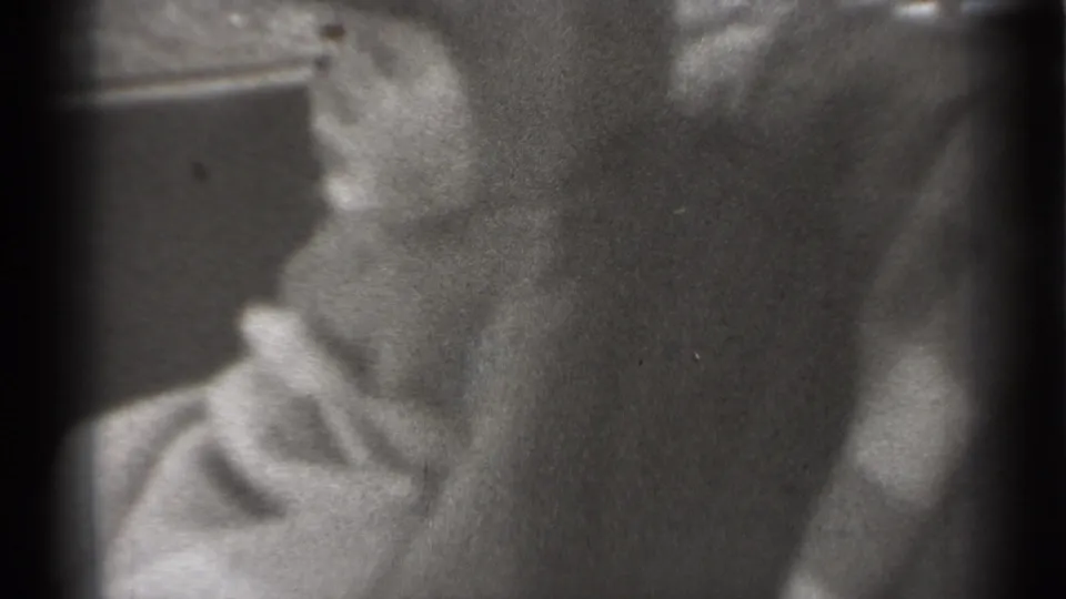 Black-and-white 8mm frame showing a baby leaning near a dark car-seat or cushion surface, with details softened by focus.