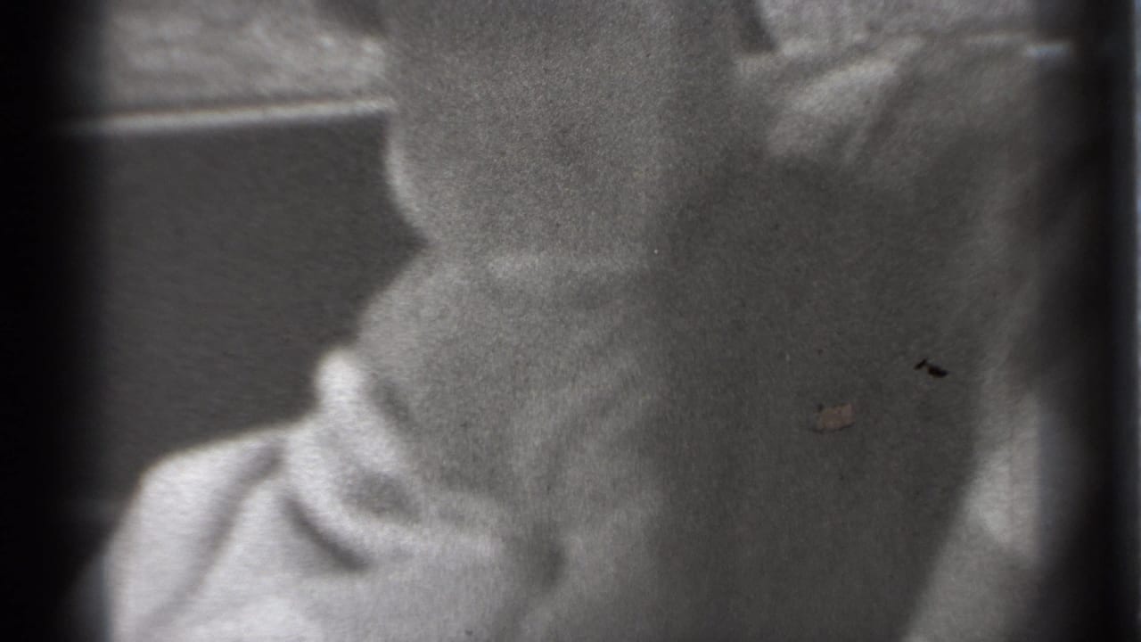 1939 New York home-movie frame showing a baby in light clothing seated inside a car, with the face partly obscured by soft focus and window framing.