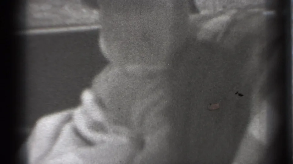 1939 New York home-movie frame showing a baby in light clothing seated inside a car, with the face partly obscured by soft focus and window framing.