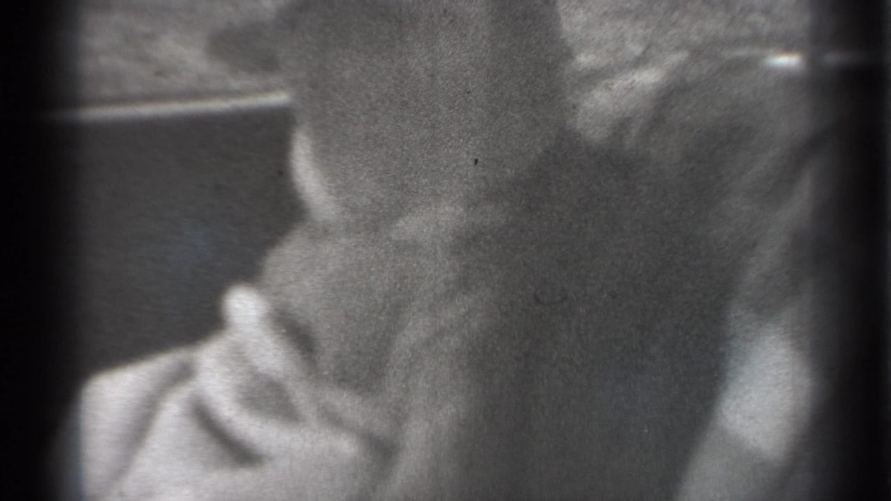 Black-and-white 8mm frame with a partial face near the left side, soft focus, heavy grain, and dark vertical film edges.