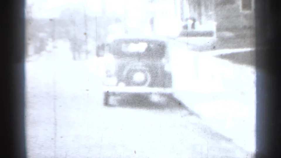 Overexposed black-and-white 8mm view of a vintage car from the rear on a New York street, with curb and building edge visible.