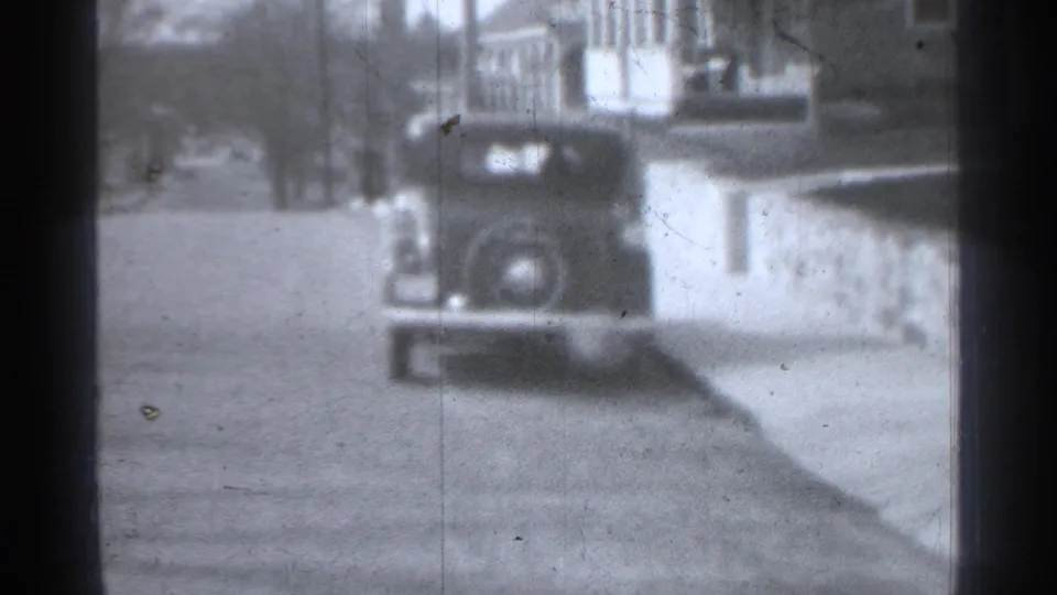 Black-and-white 8mm frame showing a rear-mounted spare tire on a vintage car along a quiet New York curbside street.