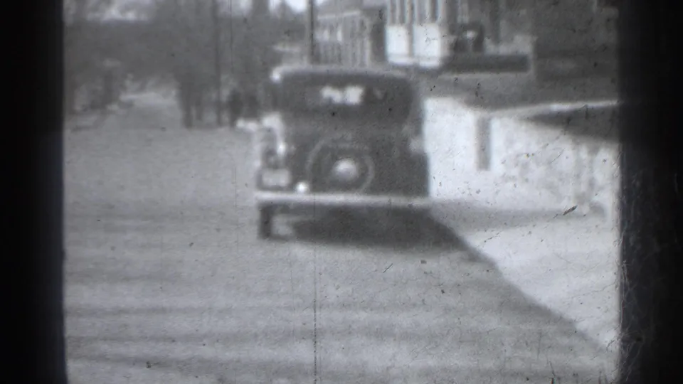 1939 New York home-movie frame showing a boxy parked car near a residential facade and sidewalk, with heavy gate edge and film wear.
