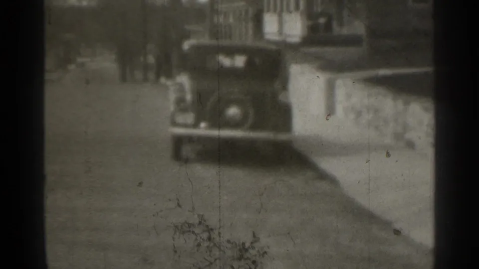 1939 New York home-movie frame holding on a parked car along a residential street, with visible dust and uneven exposure.
