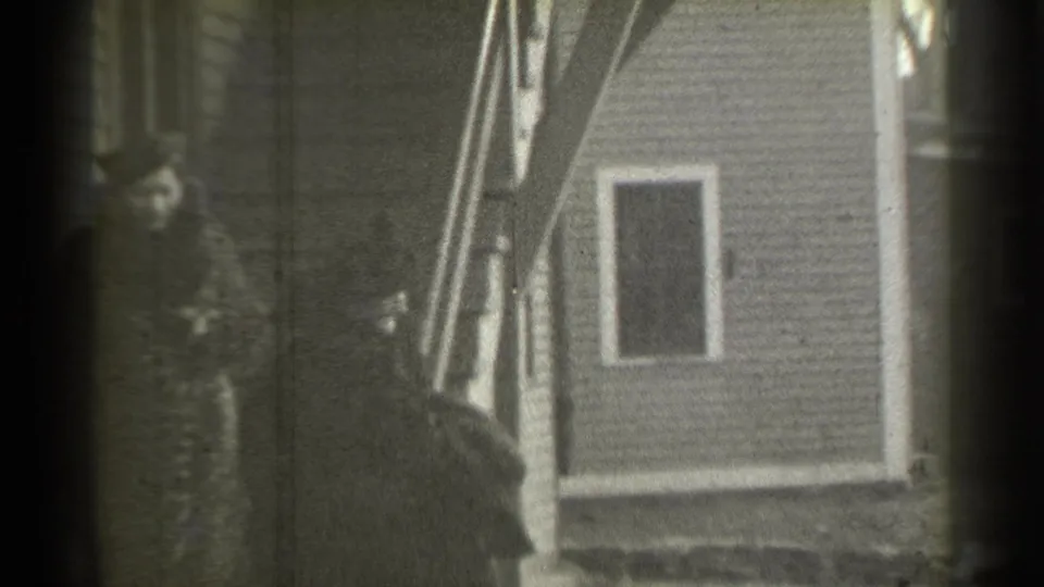 A woman in a dark fur coat and hat stands on the stairs of a New York residence, with another person partly visible below.