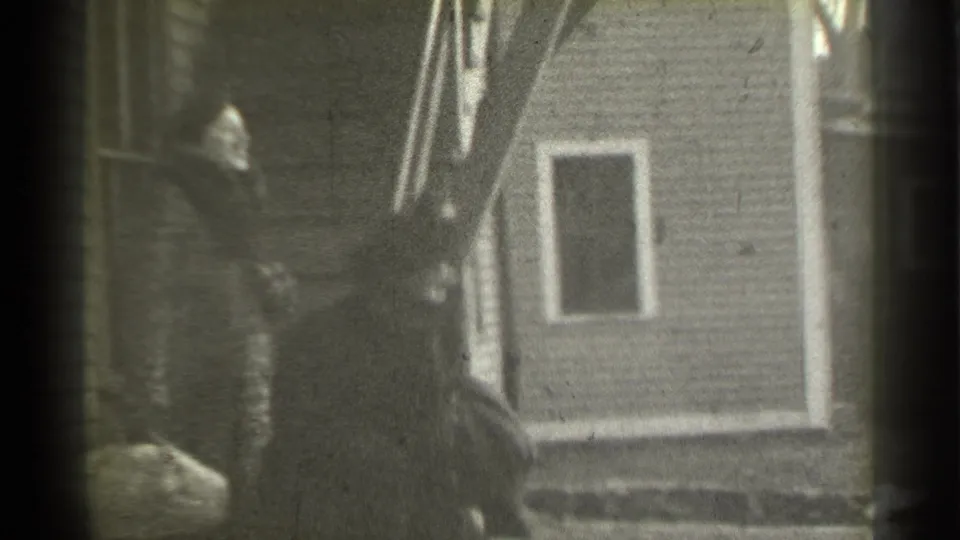 A woman in a fur coat and hat moves down exterior stairs with luggage beside a clapboard house in 1939 New York footage.