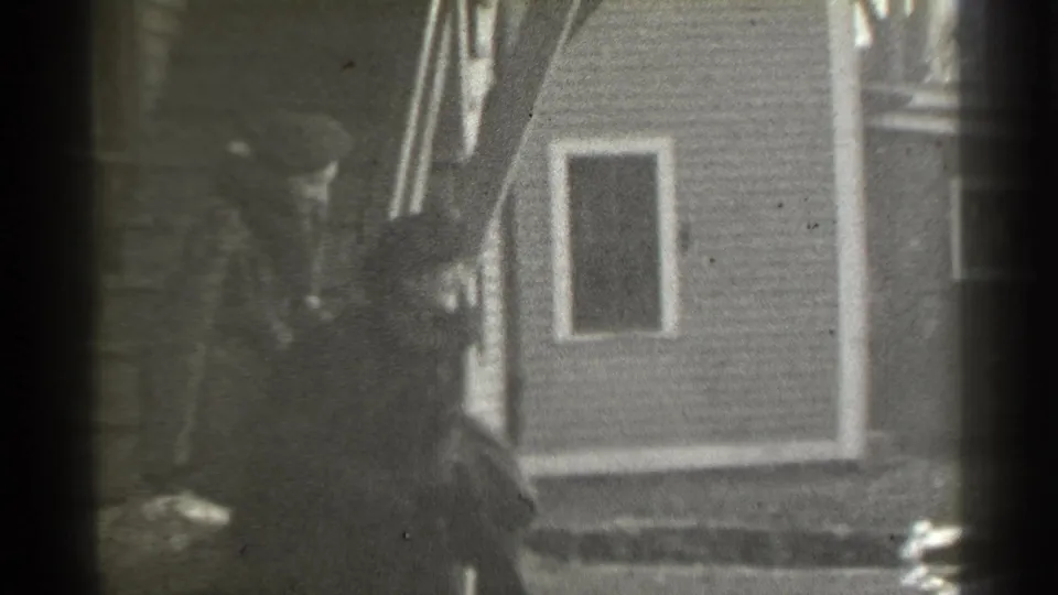 1939 New York home-movie frame showing a woman near a modest house exterior, with porch framing and soft 8mm grain.