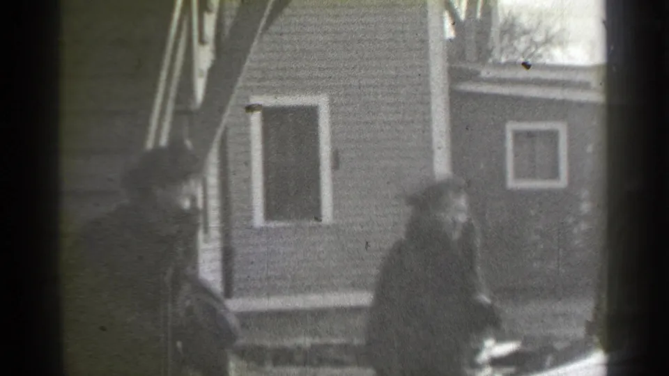 A woman in dark outerwear walks past a New York residential exterior, with a neighboring structure visible behind her.