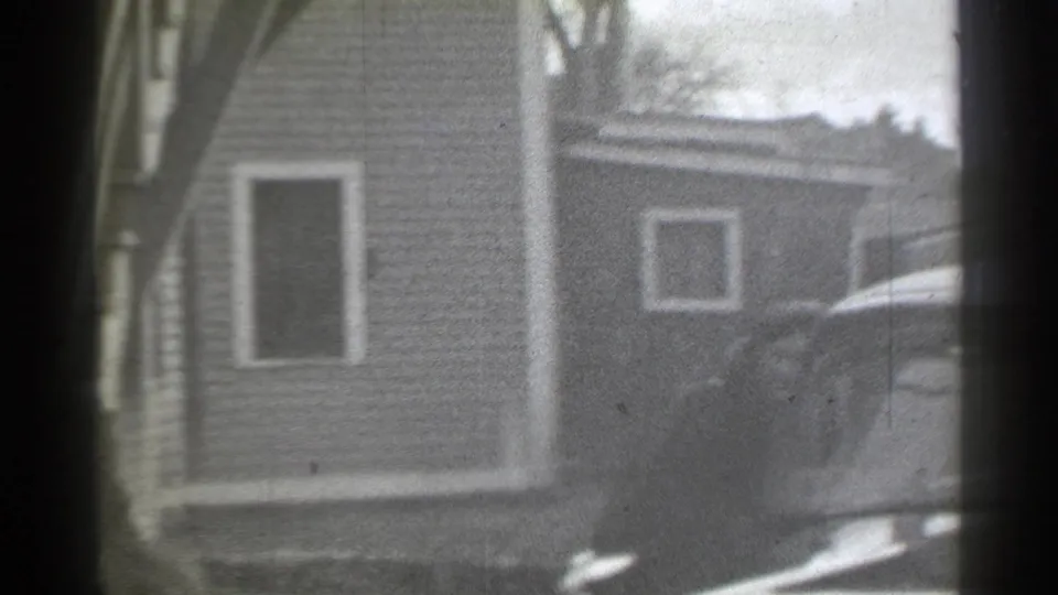 Residential New York frame with the house at left and a rounded automobile entering view as the woman nears the car.