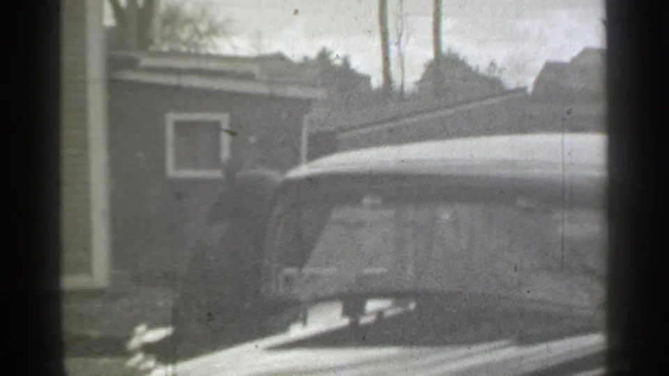 The woman is partly obscured beside a period car in a residential New York yard, with buildings and bare trees behind.