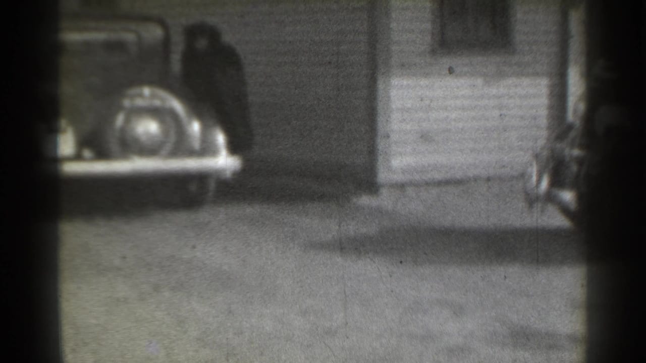 1939 New York home-movie frame showing a parked classic car near clapboard siding and a light-colored residential door.