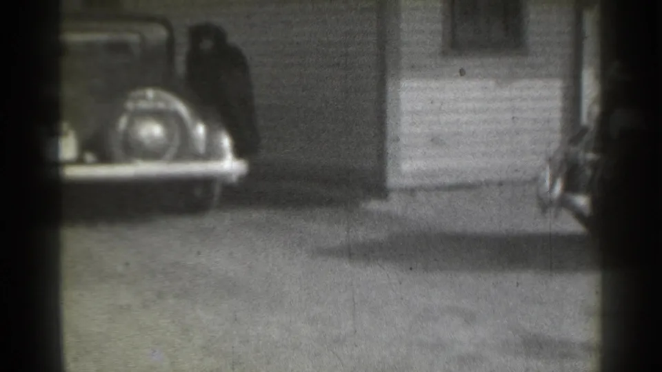 1939 New York home-movie frame showing a parked classic car near clapboard siding and a light-colored residential door.