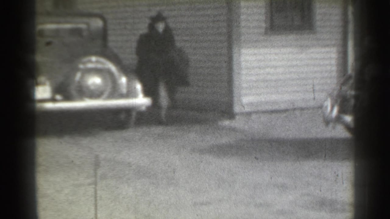 Woman in a dark coat walking near a parked classic car and house siding.