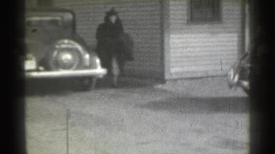 1939 New York frame with a woman in a dark coat and hat walking near a parked classic car outside a clapboard-sided building.