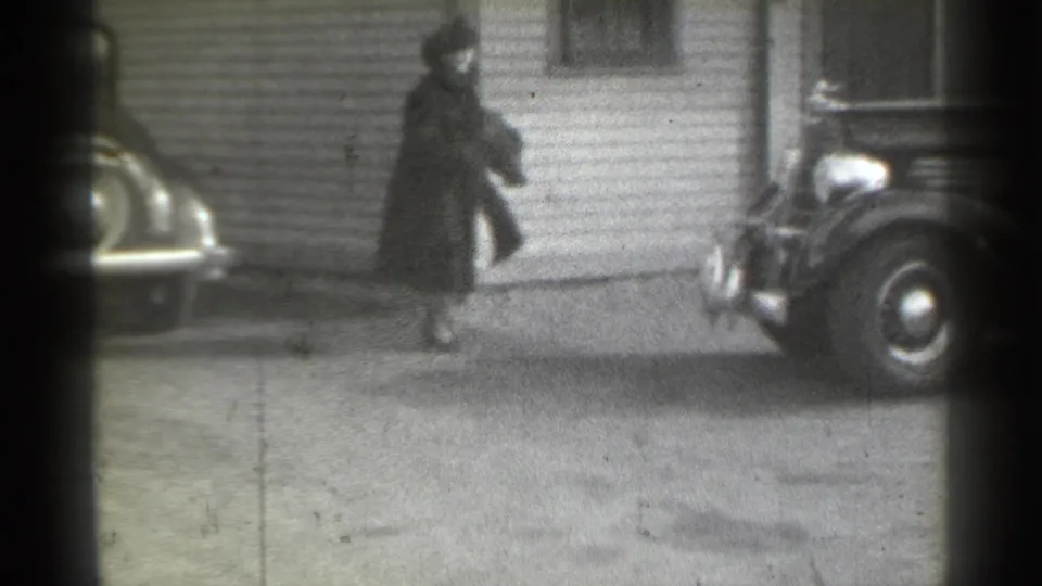1939 New York home-movie frame showing a woman in dark outerwear walking between parked classic cars in a residential driveway area.