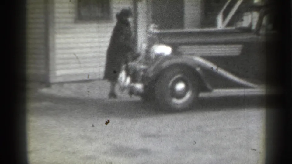 1939 New York frame showing a woman in a dark coat and hat beside the front fender and headlamp of a parked classic car.