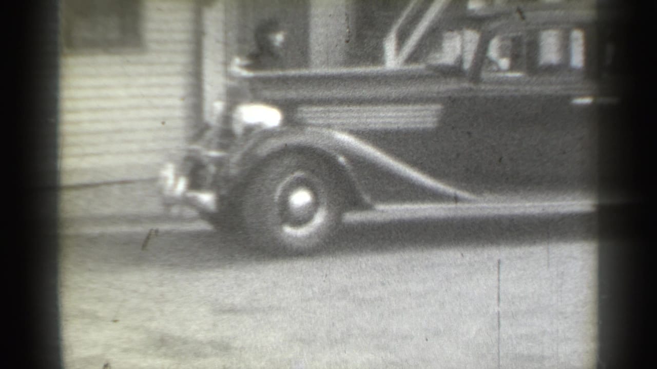 1939 New York home-movie frame showing a dark rounded sedan moving past clapboard siding, with a person briefly visible near the front.