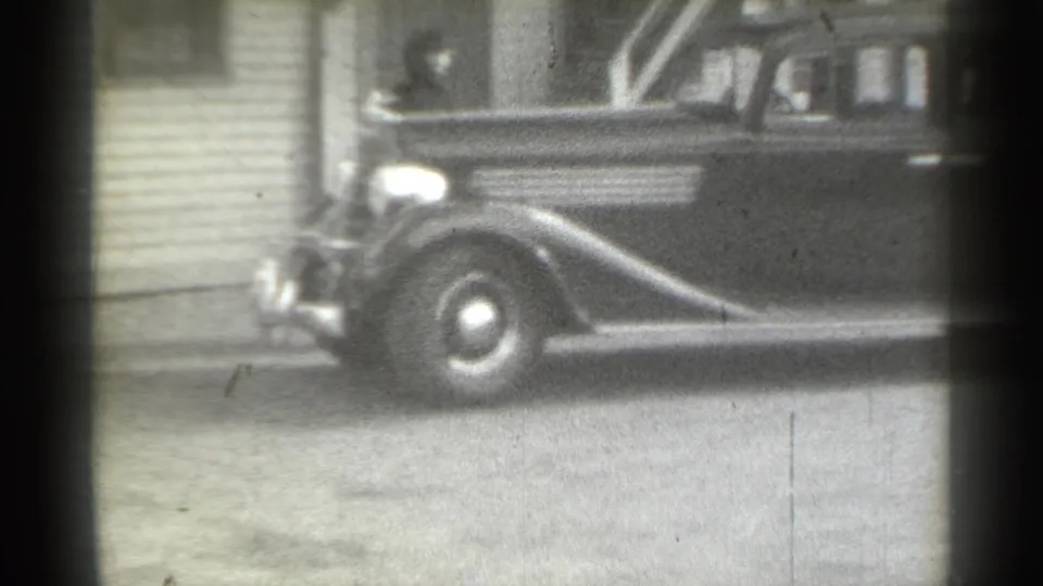 1939 New York home-movie frame showing a dark rounded sedan moving past clapboard siding, with a person briefly visible near the front.